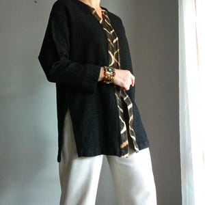 Op de afbeelding: Zwarte tuniek met een V-hals en zijsplitten. De tuniek heeft een patroonaccent aan de voorkant. Het model draagt een witte broek en een kleurrijke armband. De tuniek heeft een losse pasvorm.