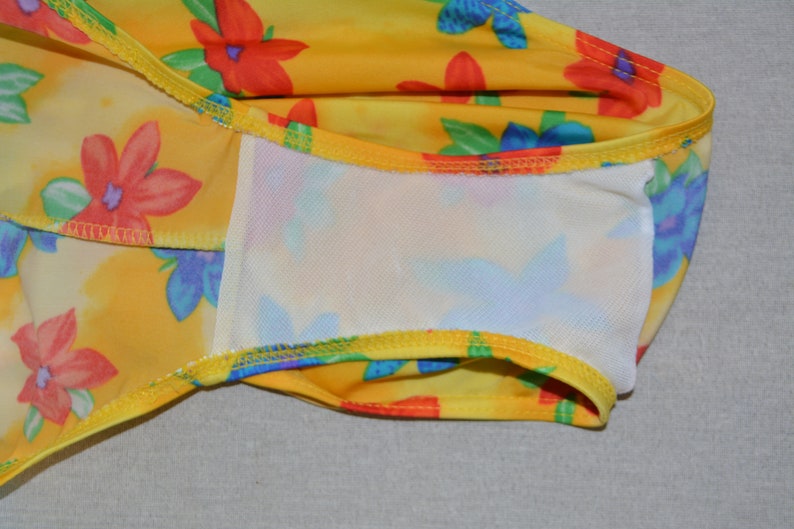 K&ouml;nnte beinhalten: Gelber Badeanzug mit Blumenmuster mit roten, blauen und gr&uuml;nen Blumen. Der Badeanzug hat ein wei&szlig;es Netzfutter.
