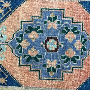 May include: A close-up of a small, rectangular rug with a central geometric design. The rug features a light pink background with a central blue design and blue borders. The design includes floral and geometric shapes in shades of blue, green, and pink.