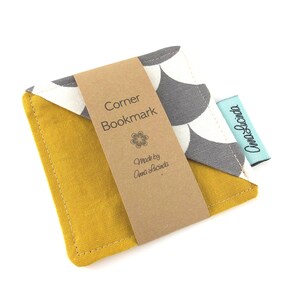 May include: A yellow and grey fabric corner bookmark with a white and grey geometric pattern. The bookmark has a brown paper tag that says "Corner Bookmark" and "Made by Anna Lucinda".