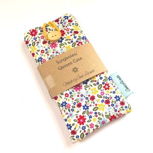 May include: A white fabric sunglasses case with a floral print in shades of red, pink, yellow, blue, and green. The case has a brown paper label that reads "Sunglasses/Glasses Case Made by Anna Lucinda" and a blue label that reads "Anna Lucinda".