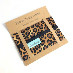 May include: A brown cardboard box with a leopard print fabric inside. The box has the text "Pocket Tissue Holder Made by Anna Lucinda" printed on the front. The fabric has a black trim and a small white label with the text "Anna Lucinda" printed on it.