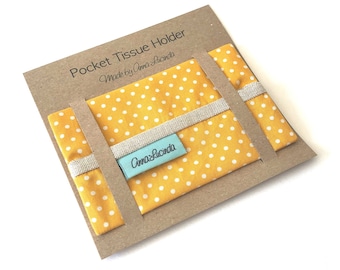 May include: A yellow fabric pocket tissue holder with white polka dots. The holder is packaged on a brown card with the text "Pocket Tissue Holder Made by Anna Lucinda" and a blue label with the text "Anna Lucinda".