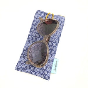 May include: A blue and white patterned glasses case with a brown and gold pair of sunglasses inside. The case has a blue and white geometric pattern and a small blue label with the text "Cannas & Lacrima" on it.