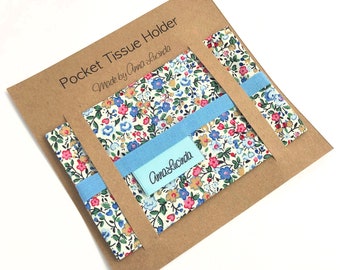 May include: A brown cardboard package containing a blue and white floral fabric tissue holder. The package is labelled "Pocket Tissue Holder Made by Anna Lucinda". The tissue holder has a blue ribbon and a label with the text "Anna Lucinda" printed on it.
