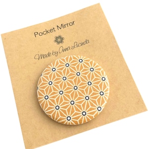 May include: A round pocket mirror with a yellow and white geometric pattern. The mirror is on a brown card with the text "Pocket Mirror" and "Made by Anna Lucinda".