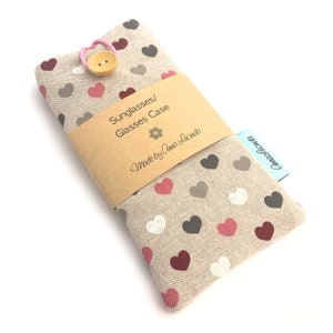 May include: A beige fabric sunglasses and glasses case featuring a repeating pattern of small hearts in shades of red, dark gray, and white. The case is secured with a light pink elastic loop and a wooden button, wrapped in a tan paper band.