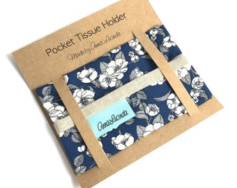 May include: A blue and white floral fabric pocket tissue holder with a beige linen trim. The holder is packaged in a brown cardboard box with the text "Pocket Tissue Holder Made by Anna Lucinda" printed on the front. The box also has a blue and white floral fabric label with the text "Anna Lucinda" printed on it.