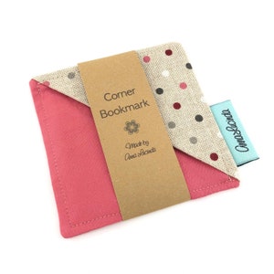 May include: A pink and beige fabric corner bookmark with a brown label that reads "Corner Bookmark" and "Made by Anna Lucinda".