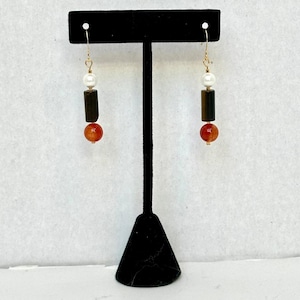 May include: A pair of dangle earrings with gold-colored hooks. Each earring features a white pearl, a dark brown rectangular bead, and a round orange bead. The earrings are displayed on a black jewelry stand against a white background.