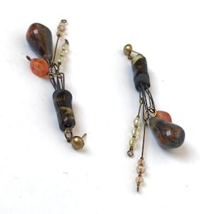 Pode incluir: Um par de brincos pendentes com contas de vidro em forma de lágrima marrom e preto, contas laranja e detalhes metálicos dourados.