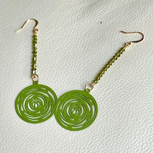May include: A pair of green earrings with a spiral design. The earrings have gold-colored ear wires and are connected to a strand of small green beads. The earrings are on a white surface.