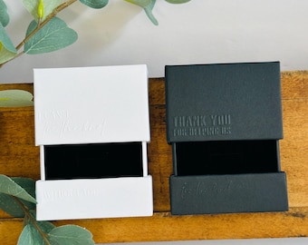 Könnte beinhalten: Zwei offene Schmuckschatullen, eine weiße und eine schwarze, stehen nebeneinander auf einer Holzoberfläche. Die weiße Schachtel trägt den Text "I CAN'T for the rest of WITHOUT YOU" und die schwarze Schachtel lautet "THANK YOU FOR HELPING US for the Pearl".