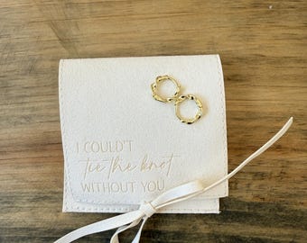 Könnte beinhalten: Ein weißer Samtbeutel mit einem goldenen Band, das zu einer Schleife gebunden ist. Der Beutel hat den Text "Ich konnte den Knoten nicht ohne dich binden" auf der Vorderseite geprägt. Zwei goldene Creolen befinden sich im Beutel.