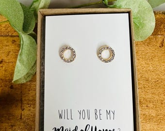 Könnte beinhalten: Ein Paar goldfarbene, kreisförmige Ohrstecker mit klaren Kristallen, präsentiert auf einer weißen Karte in einer braunen Box. Auf der Karte steht "WILL YOU BE MY Maid of Honor?" in schwarzer Schrift. Grünes Laub akzentuiert den Hintergrund.