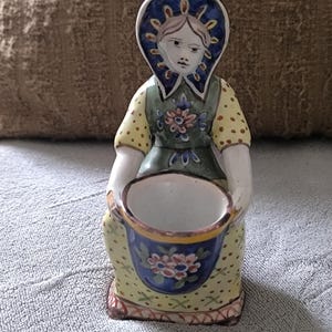 Peut inclure: Figurine en céramique d'une femme en costume traditionnel, tenant un petit seau bleu décoré de fleurs. La figurine a une robe jaune avec des accents verts et une coiffe bleue. Le visage est peint avec des traits délicats.