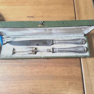 Pode incluir: Um conjunto vintage de faca e garfo de prata em uma caixa forrada de veludo verde. A faca e o garfo têm cabos ornamentados com um design decorativo. A caixa retangular tem um fecho de fecho.