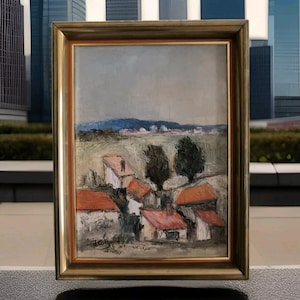 May include: A framed oil painting depicting a rural landscape with a cluster of houses with red roofs. The houses are set against a backdrop of rolling hills and a blue sky. The painting is signed "Travaglini" in the lower right corner.