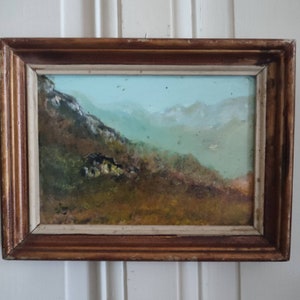 May include: A framed oil painting depicting a landscape with a mountain range in the background. The foreground features a grassy hillside with a small cluster of trees. The sky is a light blue with a few wispy clouds.