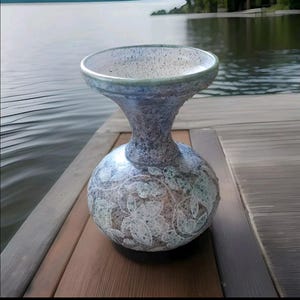 Puede incluir: Un jarrón de cerámica hecho a mano con una parte superior ancha y poco profunda y una base bulbosa. El jarrón presenta un acabado texturizado y moteado en tonos azules, grises y blancos, con un diseño floral en la base. El jarrón se exhibe sobre una superficie de madera.