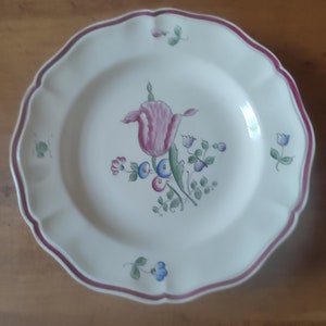 May include: A white ceramic plate with a pink and green floral design. The plate has a scalloped edge and a thin red rim.