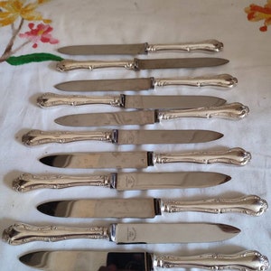 Może przedstawiać: Zestaw ośmiu srebrnych noży do masła z ozdobnymi uchwytami. Noże mają polerowane wykończenie i są ułożone w rzędzie na białym obrusie.