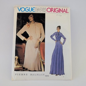 May include: A Vogue Paris Original sewing pattern envelope, number 1218, featuring a Pierre Balmain design. The cover displays a photograph of a beige long-sleeved dress with a high neckline and a matching illustration of the dress in purple.