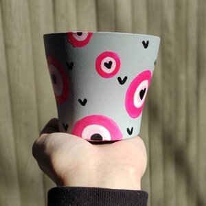 Puede incluir: Una maceta de cerámica gris con un diseño de ojo rosa y blanco y corazones negros.