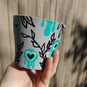 Puede incluir: Una maceta de cerámica gris con un diseño pintado a mano que presenta hojas negras, goteos de color turquesa y corazones blancos y negros.