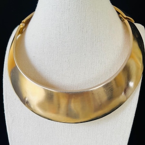 May include: A wide, gold-toned metal collar necklace with a smooth, polished finish. The piece features a curved, crescent-shaped design that sits flat against the neck, with a visible hinged clasp mechanism on the side for secure closure.