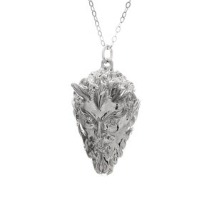 May include: A silver pendant necklace featuring a detailed, three-dimensional sculpture of a man's face with horns. The face is stylized and has a fierce expression.