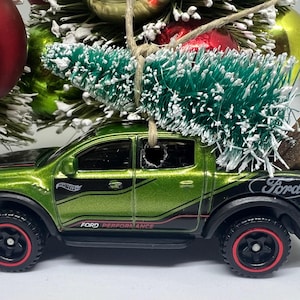 May include: A green Ford pickup truck toy with black accents and red-trimmed wheels, carrying a small green Christmas tree. The truck has the Ford logo and "Ford Performance" printed on it. A brown cylinder is in the truck bed.