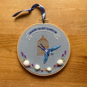 Puede incluir: Arte de bordado en aro con un pájaro azul volando fuera de una jaula dorada. El fondo es azul claro con pompones blancos y flores moradas. El texto dice "No soy un pájaro; y ninguna red me atrapa."