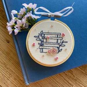 Op de afbeelding: Een kleine, ronde borduurring met een witte stoffen achtergrond. De borduurwerk toont een stapel boeken met blauwe stiksels en roze bloemen. De ring is op een blauw boek.