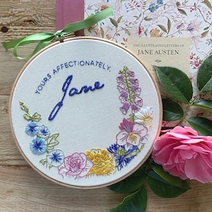 May include: A round embroidery hoop with the words "Yours Affectionately, Jane" in blue thread, surrounded by colorful embroidered flowers. A book titled "The Illustrated Letters of Jane Austen" and a pink rose are in the background.