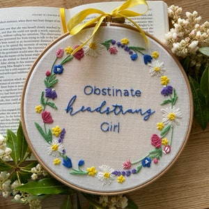 Peut inclure: Art de broderie sur un cercle avec un fond blanc et une couronne de fleurs colorées entourant le texte "Obstinate Headstrong Girl".
