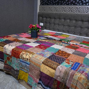 Peut inclure: Une courtepointe colorée en patchwork fabriquée à partir de tissus recyclés. La courtepointe est composée de carrés de différentes couleurs et motifs, notamment des motifs floraux, paisley et géométriques. La courtepointe est posée sur un lit avec une tête de lit grise.