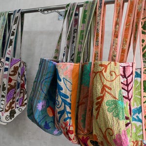 May include: A collection of colorful embroidered tote bags hanging on a metal bar. The bags feature intricate floral and geometric patterns in various colors, including blue, orange, green, and pink. The straps are also decorated with complementary designs.