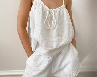 May include: White two-piece pajama set. The top has spaghetti straps and a tie detail at the neckline. The shorts have an elastic waistband and pockets. The fabric appears to be a lightweight, textured cotton.