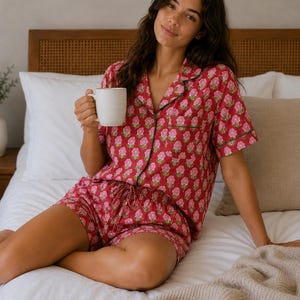 Könnte beinhalten: Ein zweiteiliger Schlafanzug bestehend aus einem kurzärmeligen Hemd mit Knopfleiste und passender Shorts aus leuchtend rotem Stoff mit einem wiederkehrenden rosa-grünen Blumenmuster, verziert mit dunkelgrünen Paspeln an Kragen und Ärmeln.