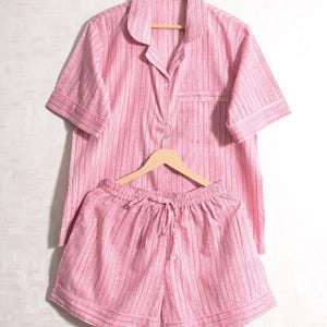 May include: A pink and white striped pajama set, including a short-sleeved button-up shirt and matching shorts. The shirt has a chest pocket and a classic collar. The shorts have a drawstring waist. Both pieces are hanging on wooden hangers against a white background.