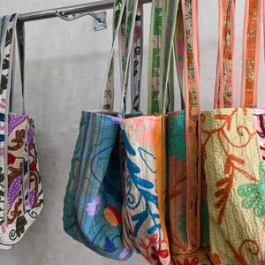 May include: A collection of colorful embroidered tote bags hanging from a metal bar. The bags feature various patterns and colors, including floral and abstract designs. The straps are also decorated with complementary patterns.
