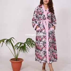 May include: A white floral print robe with pink and white stripes on the sides. The robe has a tie belt and two pockets. The model is wearing the robe and white sandals.