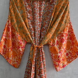 May include: An orange floral print kimono robe with wide sleeves and a tie closure. The robe features a vibrant pattern of flowers and leaves in shades of orange, red, and white. The robe is hanging on a wooden hanger.