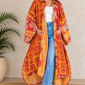 May include: A long-sleeved, button-down duster coat featuring a vibrant patchwork pattern in shades of red, orange, and yellow. The garment includes a classic collar, wide sleeves, and two large front patch pockets, worn over a white top and blue jeans.
