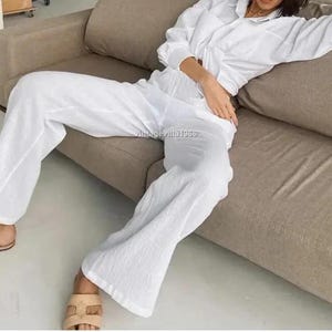 Può includere: Un completo pantalone in lino bianco con un pantalone a gamba larga e ampio. Il completo è realizzato in un tessuto leggero e traspirante e presenta una camicia abbottonata con maniche lunghe.