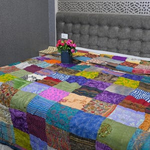 May include: A colorful patchwork quilt made from various fabrics in shades of blue, green, purple, pink, and brown. The quilt is laid out on a bed with a gray headboard.