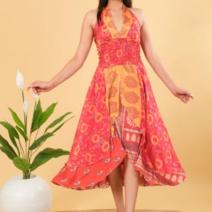 Peut inclure: Robe à col halter rouge et orange avec un motif floral. La robe a un corsage ajusté et une jupe asymétrique. Le modèle se tient devant un fond beige. La robe mesure environ 100 cm de long.