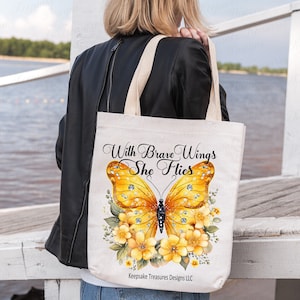 May include: A white canvas tote bag with a yellow butterfly and floral design. The text "With Brave Wings She Flies" is printed on the bag. The bag is being held by a person.
