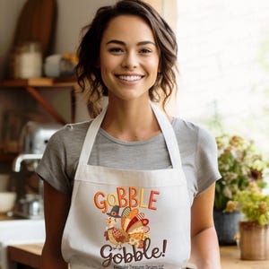 Puede incluir: Un delantal blanco con un diseño de pavo colorido y las palabras "GOBBLE GOBBLE!" en naranja y marrón. El delantal lo lleva una persona con una camiseta gris.